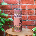 Pink tumbler with leopard print design on a wooden block against a brick wall.